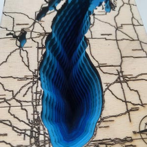Könnte beinhalten: Eine geschichtete bathymetrische Karte des Lake Michigan aus Holz mit lasergeschnittenen Konturen. Das Wasser wird durch gestapelte Blautöne von hellem Cyan bis zu tiefem Marineblau dargestellt, während das Land aus hellem Holz besteht.