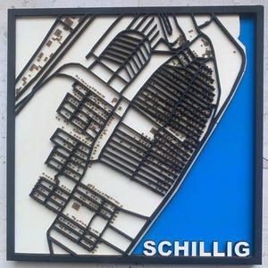 Puede incluir: Un mapa de madera enmarcado y en capas de una zona costera, con contornos negros y un fondo blanco. El mapa incluye representaciones detalladas de edificios y calles, con una sección azul que representa el mar. La palabra "SCHILLIG" se muestra en blanco.