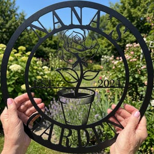 Può includere: Cartello da giardino in metallo nero con le parole "Nana's Garden" e "Est. 2024" ritagliate. Una rosa in vaso è al centro. Il cartello è tenuto davanti a un giardino.