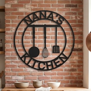 May include: Black metal kitchen sign with the words "NANA'S KITCHEN" in a circular design. The sign features silhouettes of a ladle, whisk, and spatula. The sign is mounted on a red brick wall.