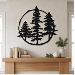 Pode incluir: Uma peça de arte de parede circular em metal preto com um design de silhueta de três pinheiros. A obra de arte é composta por uma moldura externa fina que envolve as formas detalhadas das árvores, criando um contraste com a parede.