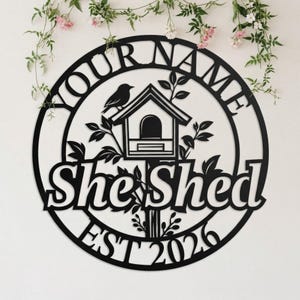 May include: Black metal sign with the words "YOUR NAME" and "She Shed" in a circular design. A bird and birdhouse are in the center, with the year "EST 2026" at the bottom. Decorative floral elements are present.