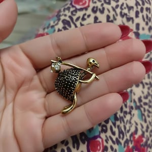May include: A gold-toned brooch in the shape of an ostrich carrying a bundle. The ostrich has a black, textured body and a detailed head. The bundle is adorned with clear crystals. The brooch is held in a hand.