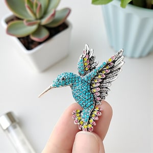 Może przedstawiać: Broszka w kształcie kolibra ze srebrnym metalowym korpusem i skrzydłami. Ciało ptaka pokryte jest turkusowymi kryształkami, z akcentami różu, żółci i błękitu na skrzydłach i ogonie. Ozdobny dodatek.