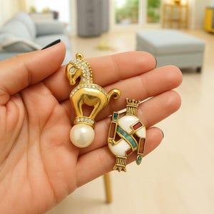 May include: Two gold-coloured brooches. One is a horse with rhinestone accents and a pearl base. The other is a white, red, and turquoise enamel design with a crown. Both are ornate and likely vintage.