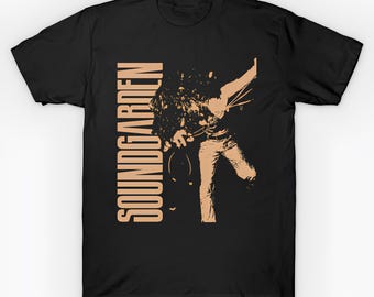 May include: Black t-shirt featuring a tan graphic of a person playing a guitar, with the band name "SOUNDGARDEN" printed vertically in tan. The design is a stylized illustration, suitable for music fans.