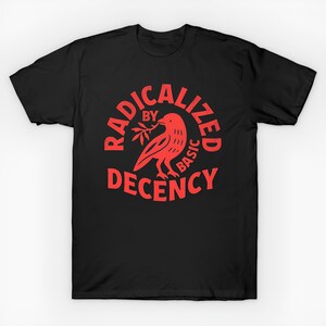 Puede incluir: Camiseta negra con un gráfico rojo. El gráfico presenta un pájaro sosteniendo una ramita y las palabras "RADICALIZED BY BASIC DECENCY" en un diseño circular. La camiseta es de cuello redondo.