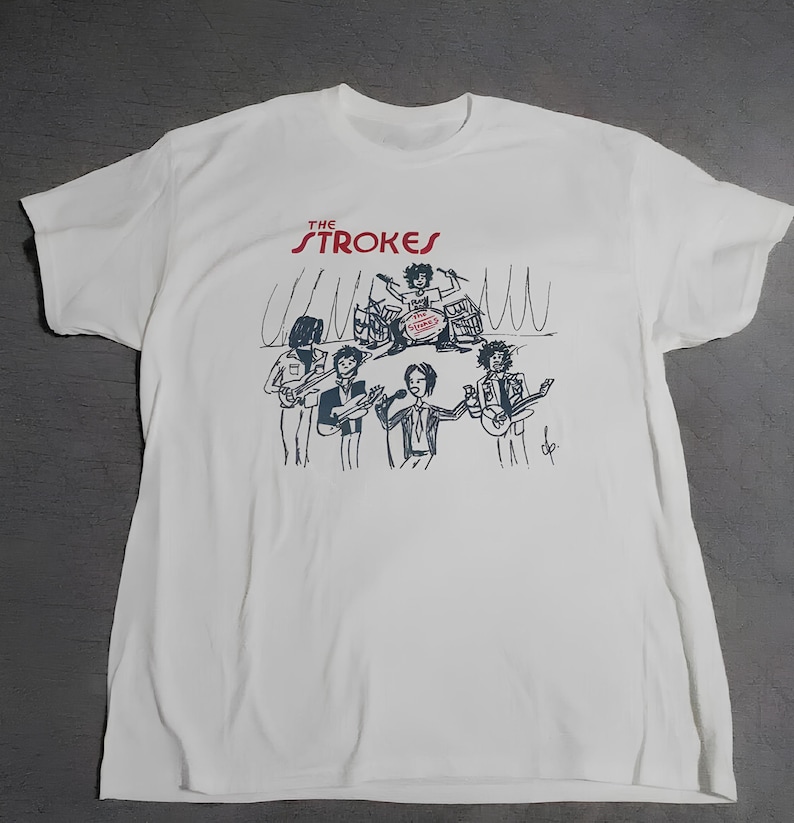 Puede incluir: Camiseta blanca con una ilustraci&oacute;n de una banda. El nombre de la banda "The Strokes" est&aacute; impreso en rojo encima de la ilustraci&oacute;n. La ilustraci&oacute;n representa una banda con un bater&iacute;a, guitarristas y un cantante. El dise&ntilde;o es en blanco y negro.
