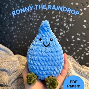 Può includere: Un peluche a forma di goccia di pioggia blu all'uncinetto, con un volto sorridente e piedini verdi. Il testo "RONNY THE RAINDROP" è sopra il peluche. Un cerchio blu con il testo "PDF Pattern" e "wooliebambooie" è in basso a destra.