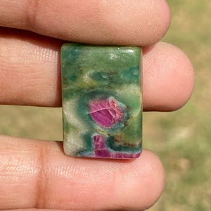 Puede incluir: Una gema rectangular con una vibrante mezcla de colores. La piedra presenta una base verde con patrones arremolinados y un área prominente de tonos rosa intenso y rojo. La gema se sostiene entre los dedos, mostrando su superficie pulida.