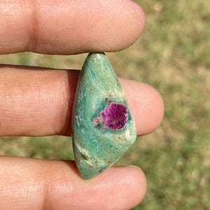 Op de afbeelding: Een gepolijste edelsteen met een teal basiskleur en een levendige rode insluitsel. De steen wordt tussen de vingers gehouden, wat de unieke vorm en natuurlijke schoonheid laat zien. De edelsteen is ongeveer 2,5 cm lang.