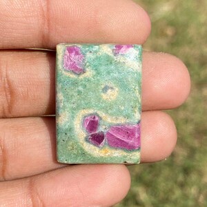 Op de afbeelding: Een rechthoekige edelsteen met een groene basiskleur en levendige roze insluitsels. De steen wordt in een hand gehouden en toont zijn gepolijste oppervlak en natuurlijke patronen. De edelsteen is ongeveer 2,5 cm lang.