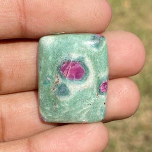 May include: A rectangular gemstone with a green base color and vibrant pink inclusions. The stone has a polished surface and is held in a hand, showcasing its natural beauty. The gemstone's unique color combination makes it a striking piece.