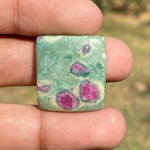 Op de afbeelding: Een vierkante edelsteen met een groene basiskleur en levendige roze en rode insluitsels. De steen wordt in een hand gehouden, waarbij het gepolijste oppervlak en de natuurlijke patronen worden getoond. De edelsteen is een uniek sieraad.