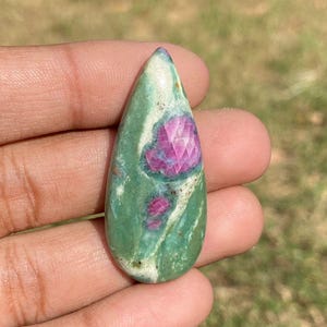 Op de afbeelding: Een druppelvormige, gepolijste edelsteen. De steen heeft een groene basis met witte adering en een centrale, gefacetteerde robijninsluiting. De robijn heeft een diepe roze kleur, die contrasteert met het groen.