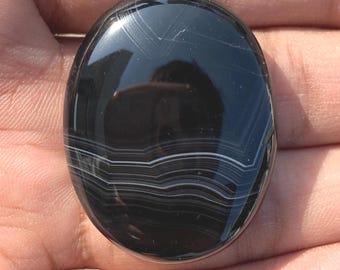 May include: A polished, oval-shaped black gemstone with white banding. The stone has a glossy surface and reflects light, showcasing its natural patterns. The gemstone is held in a hand, highlighting its size and shape.