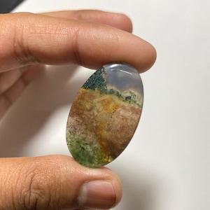May include: An oval-shaped moss agate gemstone with a landscape-like pattern. The stone features a mix of colors, including green, brown, yellow, and a touch of blue. The polished surface of the stone is smooth and reflective.