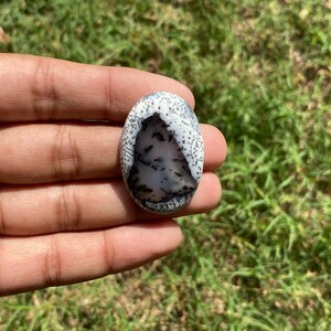 May include: An oval gemstone with a white background and detailed black and grey patterns, reminiscent of a landscape. The stone is held in a hand, highlighting its polished surface and natural beauty. A unique piece of jewellery.