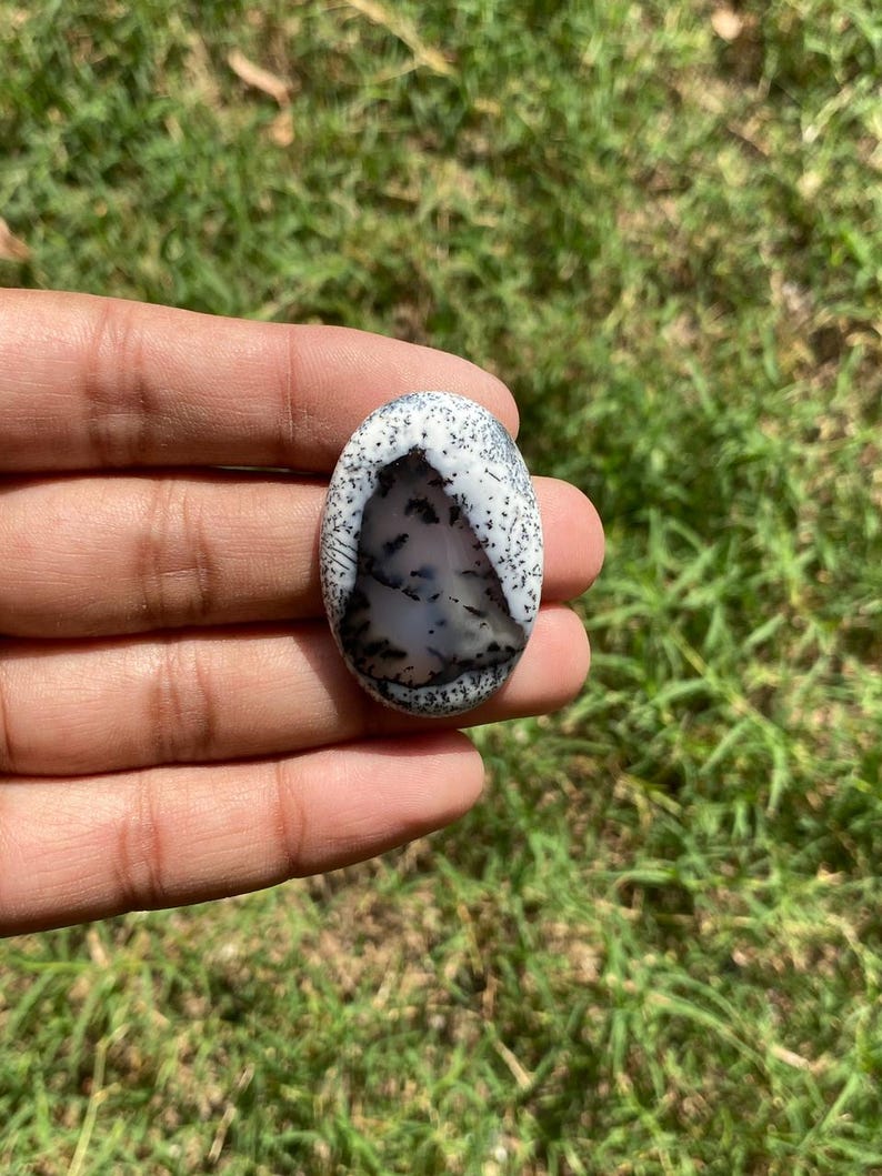 May include: An oval gemstone with a white background and detailed black and grey patterns, reminiscent of a landscape. The stone is held in a hand, highlighting its polished surface and natural beauty. A unique piece of jewellery.