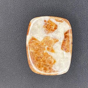 May include: A rectangular, polished gemstone with a creamy white base and embedded orange-brown crystalline structures. The stone has rounded edges and a smooth, glossy surface, showcasing natural patterns.
