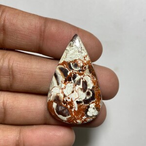 May include: A teardrop-shaped gemstone with a unique pattern. The stone features a mix of white, brown, and reddish-brown colors. The surface has a cracked appearance, adding to its natural beauty. The gemstone is held in a hand.