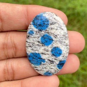 May include: Oval-shaped K2 stone with a white and gray speckled background and vibrant blue spots. The polished gemstone is held in a hand, showcasing its natural patterns and colors. The stone is approximately 2 inches long.