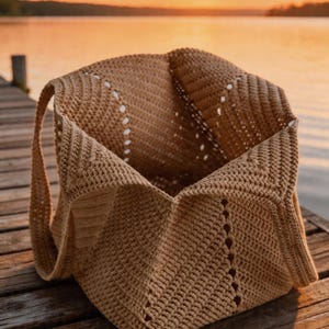 May include: A tan crochet tote bag with a rectangular base and open top. The bag has two handles and decorative openwork along the sides. The bag is sitting on a wooden dock with a lake and sunset in the background.