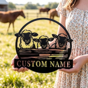 May include: Black metal sign depicting three sheep over a fence, framed in a circle. The sign reads "CUSTOM NAME" on a rectangular base. The background shows a grassy field and horses.