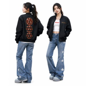 May include: A black bomber jacket with a red circular graphic on the back featuring white bird silhouettes. The front displays gold text and a white crop top with three red hearts. Blue denim jeans feature white star patterns on the legs.