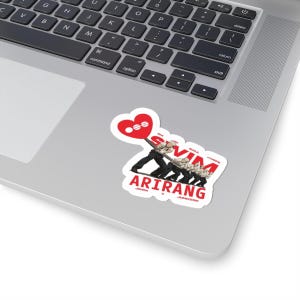 May include: A white sticker with a design of a group of people holding a heart-shaped balloon. The sticker has the text "ARIRANG" and the names of the group members. The sticker is placed on a laptop.