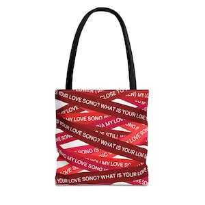 Può includere: Una borsa tote bianca con manico nero. Il design della borsa presenta nastri rossi con la scritta "WHAT IS YOUR LOVE SONG?" e altre frasi. La borsa è un accessorio elegante.