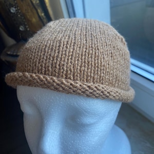 Könnte beinhalten: Eine hellbraune Strickmütze mit gerolltem Rand. Die Mütze wird auf einem weißen Schaufensterkopf präsentiert. Die Mütze ist aus weichem, strukturiertem Garn gefertigt und hat ein enges Strickmuster.
