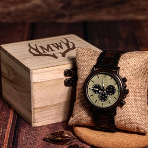 May include: A wooden watch with a black band and a light green face, displayed on a burlap pillow. A wooden box with the initials "MW" and antlers is next to the watch. The watch has three sub-dials.