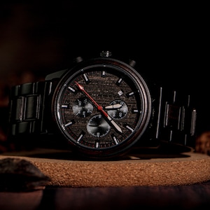 May include: A dark wooden wristwatch with a black metal band. The watch face features a wood grain pattern with black subdials and white and red hands. The watch is displayed on a brown surface.