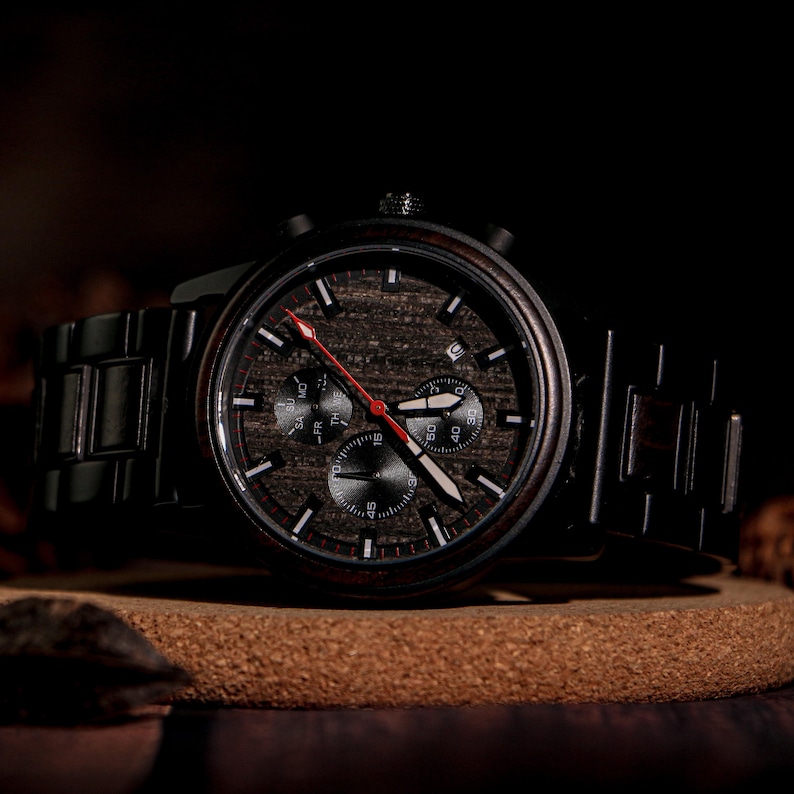 May include: A dark wooden wristwatch with a black metal band. The watch face features a wood grain pattern with black subdials and white and red hands. The watch is displayed on a brown surface.