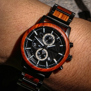 May include: A black wristwatch with a wooden bezel and band is displayed. The watch face is black with white subdials and hands. The wooden elements have a reddish-brown hue, and the watch is on a wrist.