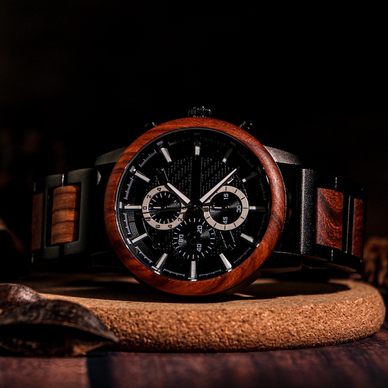 May include: A wooden wristwatch with a black face and a dark brown wooden bezel and band. The watch has three sub-dials and white hands and markers. The watch is displayed on a round, brown surface.