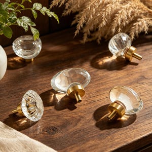 Può includere: Pomelli per mobili in vetro trasparente con basi in metallo dorato. I pomelli hanno varie forme sfaccettate e ovali. I pomelli sono esposti su una superficie di legno, con un vaso di verde e erba ornamentale secca sullo sfondo.