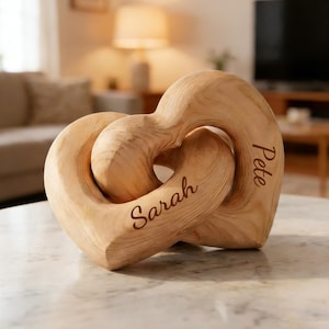 Puede incluir: Escultura de madera con dos corazones entrelazados. La madera, de color marrón claro, muestra vetas visibles. Los nombres "Sarah" y "Pete" están grabados en los corazones. Objeto decorativo.