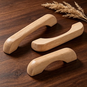 May include: Three wooden cabinet handles with a natural finish are arranged on a dark wood surface. The handles have a curved design, with a rounded grip and a slightly angled base. The wood grain is visible, adding to the rustic aesthetic.