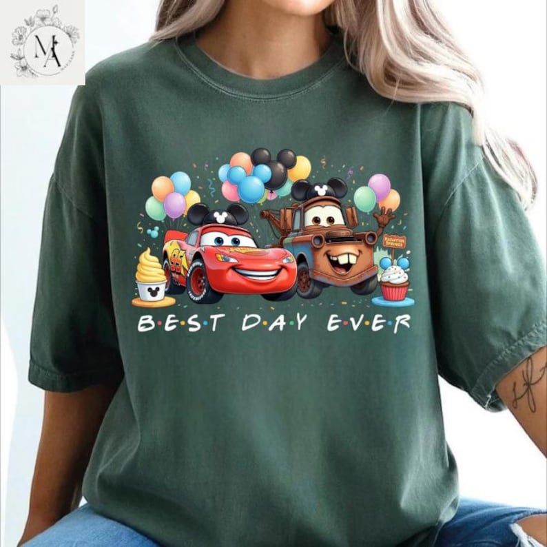 A green t-shirt with a graphic design featuring two cartoon cars, one red and one brown, surrounded by colorful balloons and a cupcake. The text "BEST DAY EVER" is written below the image.
