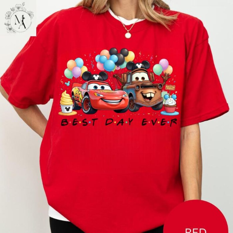 A red t-shirt with a graphic design featuring two cartoon cars, one red and one brown, surrounded by balloons and a cake. The text "BEST DAY EVER" is written below the image.