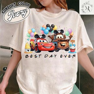 A person wearing a white t-shirt with a graphic design featuring characters from the Disney movie Cars, including Lightning McQueen, Lightning McQueen's car, and a cake.
