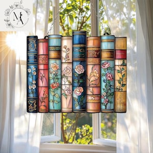 May include: Stained glass window panel in the shape of a stack of books. Each book is a different color, with floral designs and gold accents. The panel hangs from a chain in front of a window with white curtains.