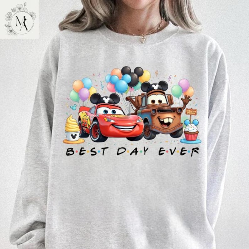 A person wearing a gray sweatshirt with a graphic design featuring two cartoon cars, one red and one brown, surrounded by various Disney-themed elements such as balloons, a cupcake, and a sign that says "Best Day Ever".