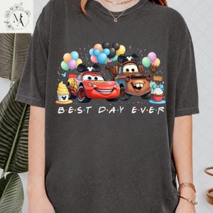 A person wearing a black t-shirt with a graphic design featuring two cartoon cars, one red and one brown, surrounded by various party elements such as balloons, cupcakes, and a Mickey Mouse hat.