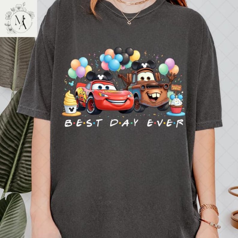 A person wearing a black t-shirt with a graphic design featuring two cartoon cars, one red and one brown, surrounded by various party elements such as balloons, cupcakes, and a Mickey Mouse hat.