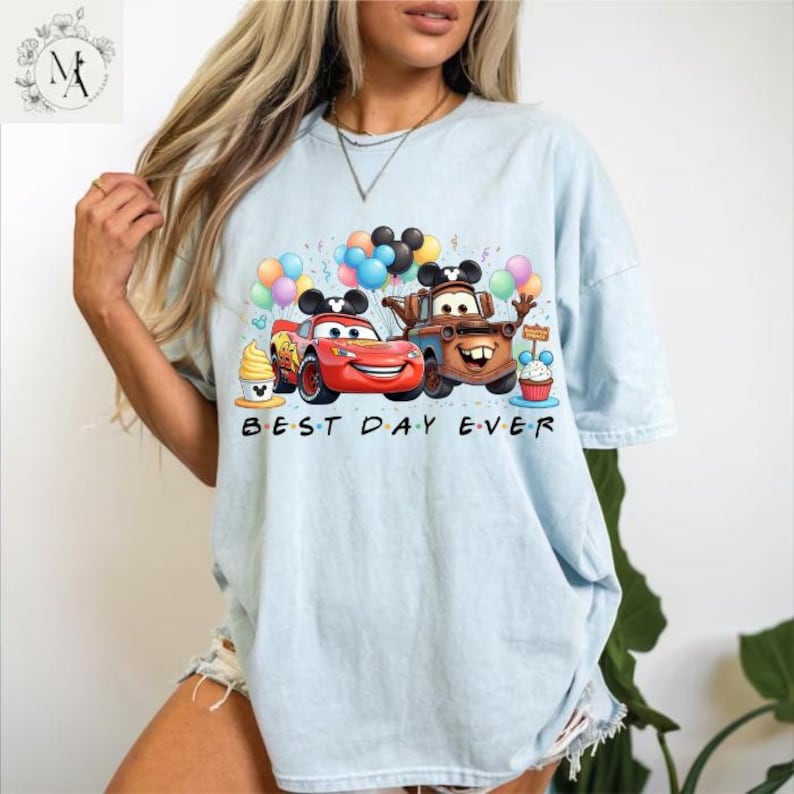 A woman wearing a light blue t-shirt with a graphic design featuring cartoon cars and balloons, along with the text "Best Day Ever".