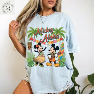 May include: Light blue t-shirt featuring Mickey and Minnie Mouse in Hawaiian attire with the text "Mickey Aloha." The graphic includes palm trees, flowers, a surfboard, and a tiki. The shirt is a relaxed fit.