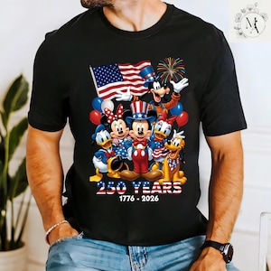 A black t-shirt with a graphic design featuring Disney characters Mickey Mouse, Minnie Mouse, Donald Duck, Goofy, and Pluto, along with an American flag and fireworks. The text below the image reads "250 YEARS 1776-2026".